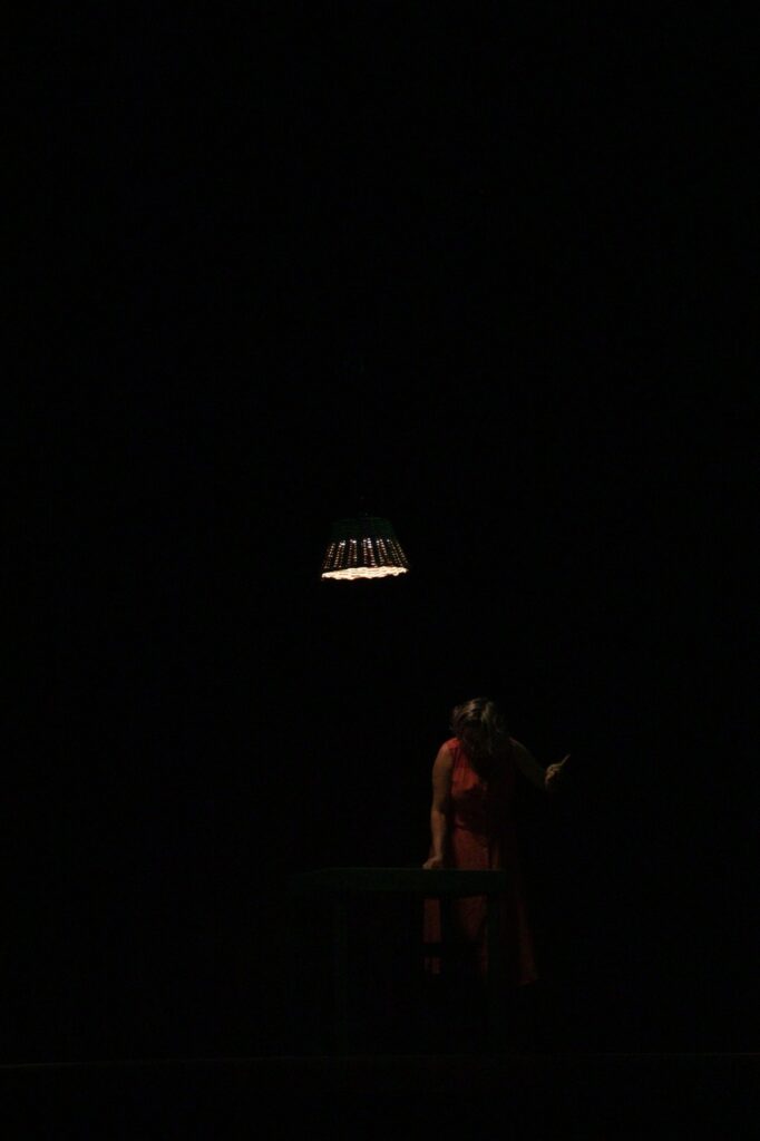 Bailarina em vestido vermelho, em cena com lâmpada pendente e fundo escuro.