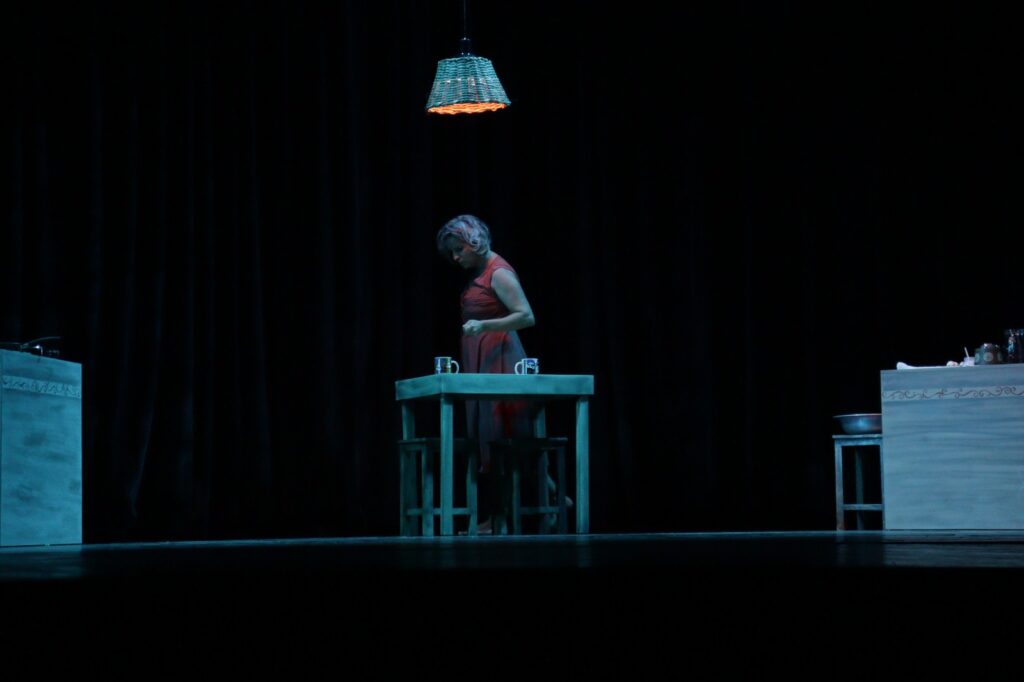 Bailarina em vestido vermelho, em cena com lâmpada pendente e fundo escuro.