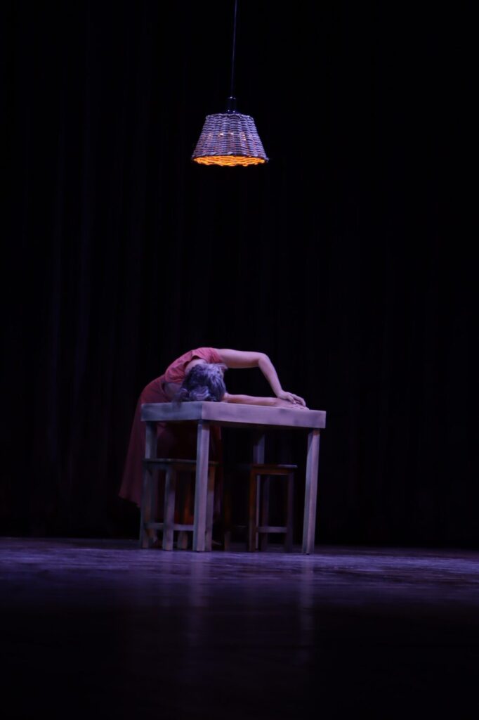 Bailarina em vestido vermelho, em cena com lâmpada pendente e fundo escuro.