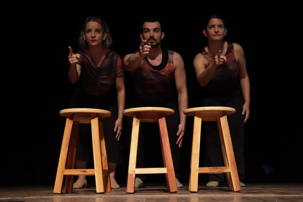 Três bailarinos em frente a banquetas, com fundo preto e iluminação cênica.