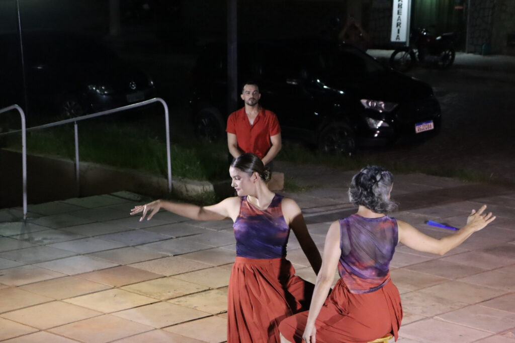 Dois bailarinos dançando em espaço externo à noite, com espectador ao fundo.