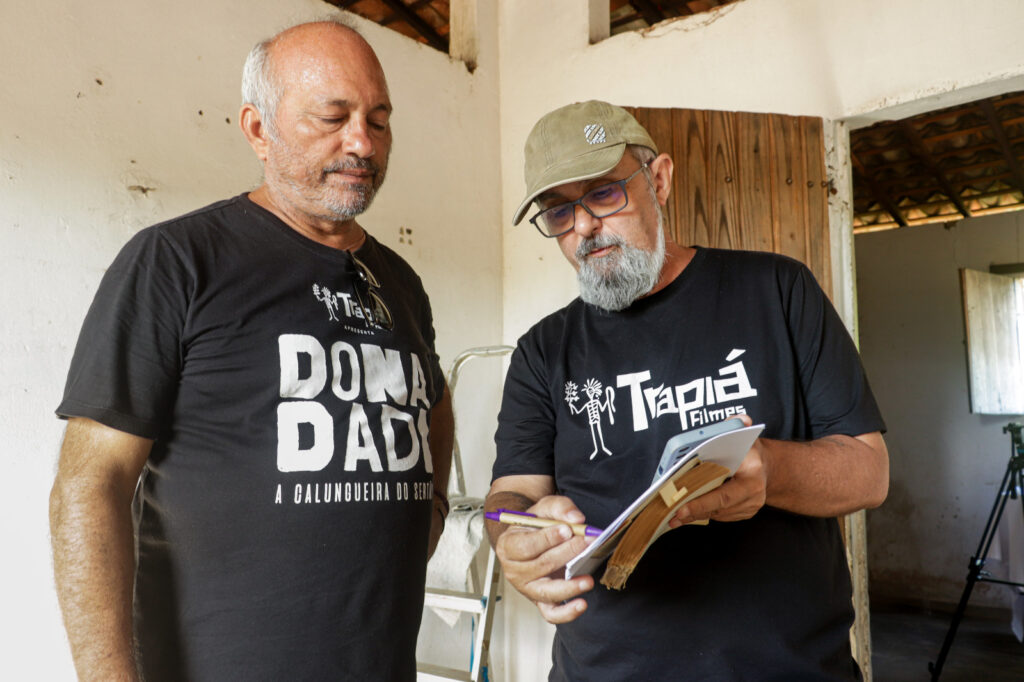 O diretor de boné e óculos revisa anotações em um caderno com outro homem em uma locação interna durante uma pausa na filmagem.