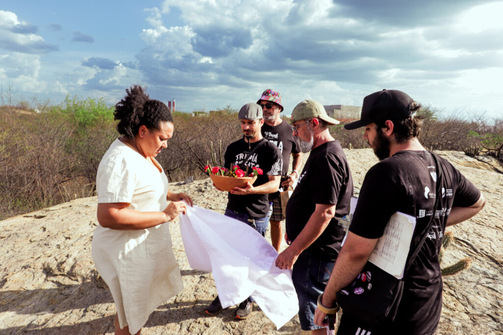 Equipe de produção e elenco reunidos em um platô rochoso, preparando um cesto com flores para a filmagem de uma cena ao ar livre.