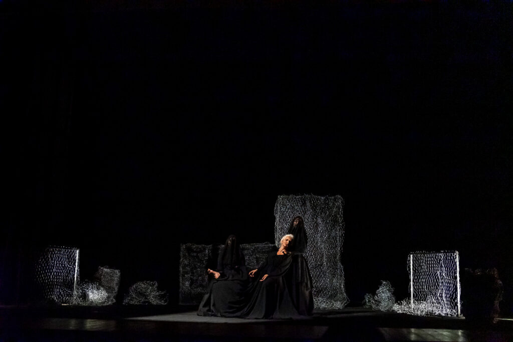 Em um palco escuro, três figuras vestidas de preto estão em cena. Uma mulher de cabelos brancos senta-se, amparada por outra. Ao fundo, painéis de rede cinza compõem o cenário.