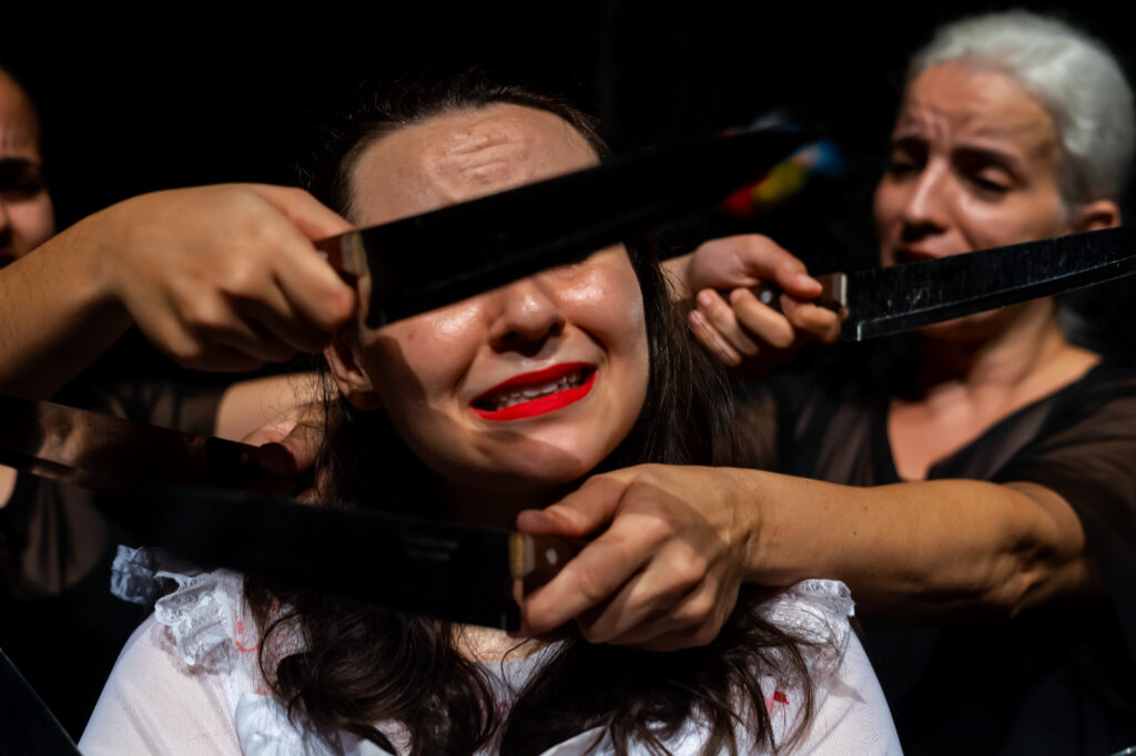 Close-up de uma mulher com expressão de angústia e batom vermelho. Mãos seguram facas apontadas para seu rosto e pescoço, em uma cena de grande tensão.